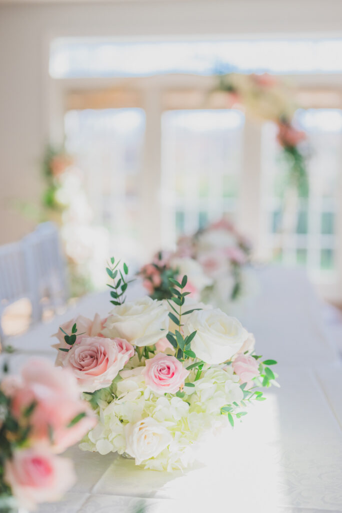 Dining table centerpiece with pink and white roses, featuring a blurred floral arch in the background, at a party in Boston.