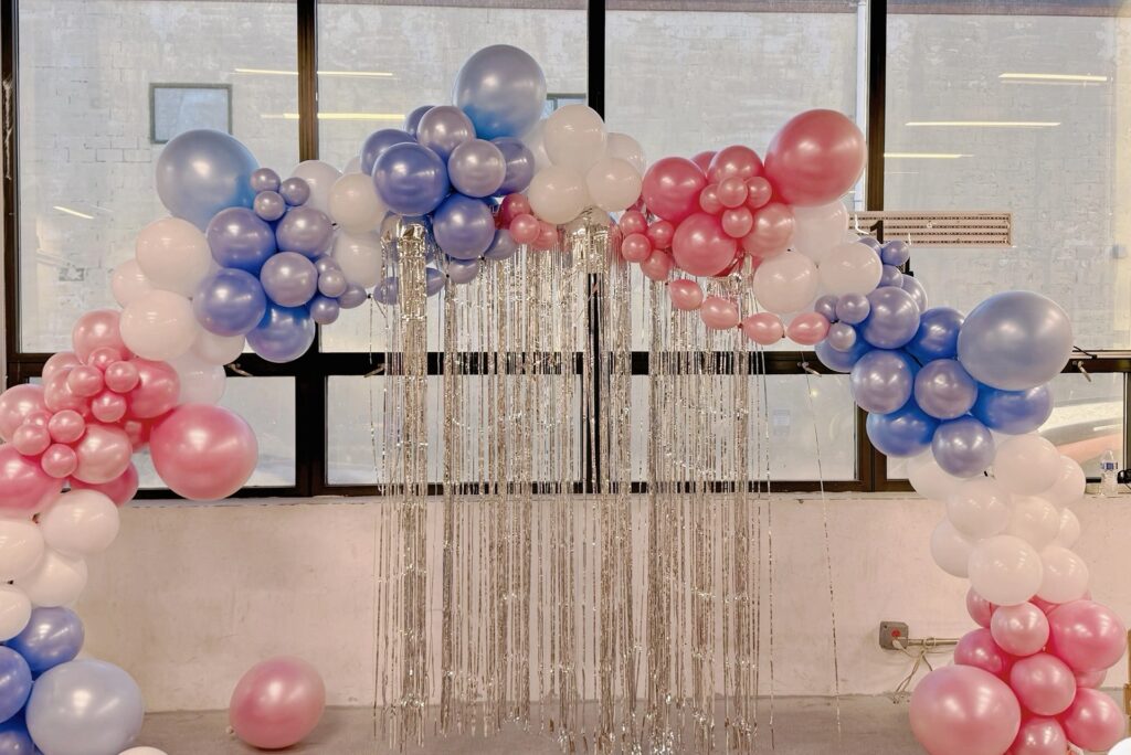 Large balloon arch decoration in pink, blue, and white balloons with silver fringe at a party in Boston.