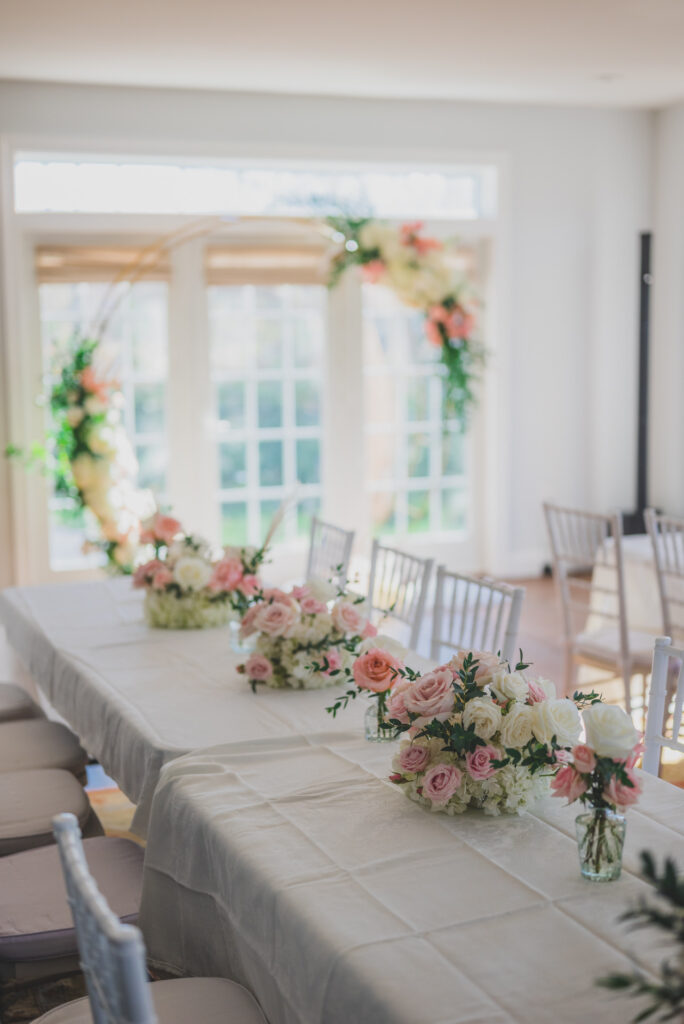 Wedding reception long tables with pink and white floral centerpieces and a gold floral arch in a bright room.